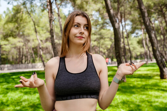 Beautiful brunette woman wearing sports bra standing on city park, outdoors clueless and confused with open arms, shrugging shoulders, no idea and doubtful face.