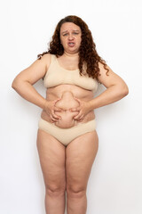 Portrait of frowning overweight woman with long curly hair wearing beige underwear, squeezing...