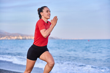 young athletic woman in a red shirt and braid running on the shore of the beach with mountains in the background
