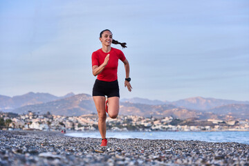 young athletic woman in a red shirt and braid running on the shore of the beach with mountains in the background - obrazy, fototapety, plakaty