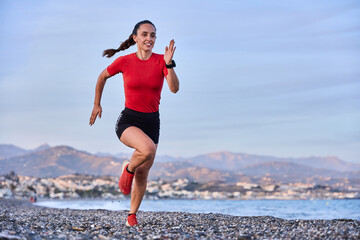 young athletic woman in a red shirt and braid running on the shore of the beach with mountains in the background