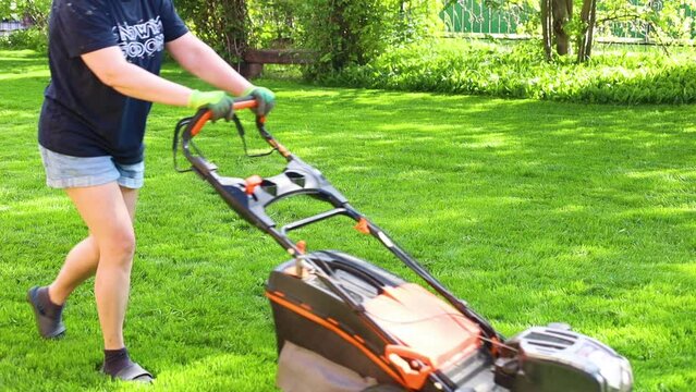 Woman mowing green lawn in backyard moving along area with lawn mower on summer sunny day closeup, slow motion. Landlady cleaning up territory, nature background. Care and maintenance of home garden.