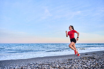young athletic woman in a red shirt and braid running on the shore of the beach with mountains in...