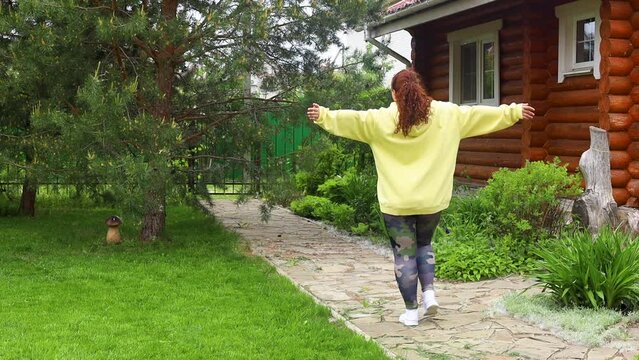 Rearview of red-haired curly fat woman with excess weight in yellow hoodie walking calm on road with raising hands in green backyard outdoor. Nature, meadow and house on background. Body positive