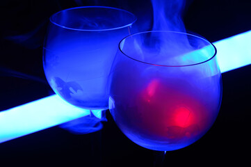 Two smoky cocktails with a neon tube, on a dark background. One of the glasses slowly igniting alcohol inside. Dangerous liquor mixture - close up photo. Background picture.