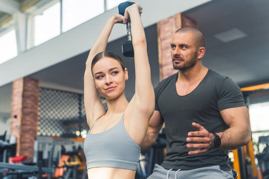 Muscular caucasian male personal trainer teaches a fit skinny caucasian brunette woman hot to properly exercise with a dumbbell. High quality photo