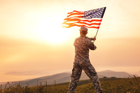 Male Soldier In The Uniform Of The American Army Waving The US Flag On Top Of A Mountain In A Clearing At Sunset