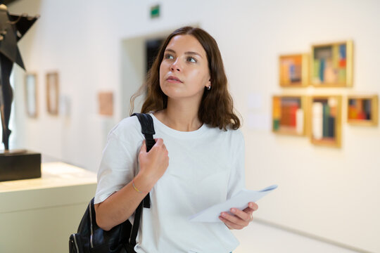 Focused girl visitor with a booklet in her hands examines an exhibit standing in an art gallery
