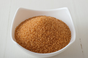 Granulated cane sugar in porcelain bowl on a white wooden table. Closeup, no people.