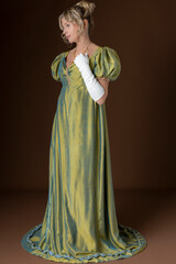 A blonde Regency woman wearing a green shot silk dress and standing against a plain backdrop