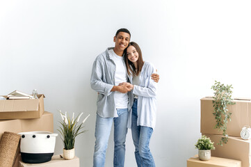 Stylish a guy and a girl in love hugging each other, multiracial excited couple standing near a white wall, between cardboard boxes, in their new apartment, looking at camera, smiling happily
