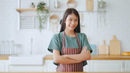 Happy young asian woman wearing apron and standing in kitchen room. Beautiful female smiling and looking at camera feeling confident