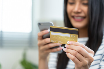 Asian attractive woman enjoy shopping online in living room at home. Beautiful girl sit on sofa, holding credit card to make e bank online payment after use phone purchase goods in web store in house.