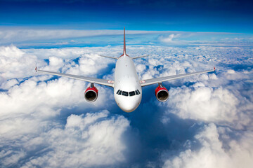 White passenger plane with red engines in flight. Aircraft flies high in the blue sky above the clouds. Front view.