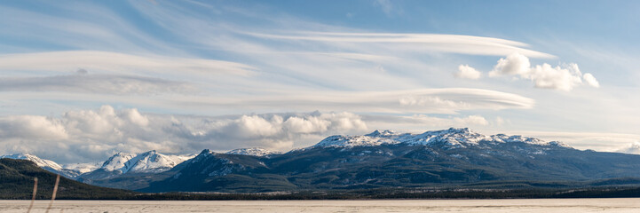 Panoramic shot of snow capped mountain view scenery seen in northern British Columbia during spring time with blue sky day and clouds.