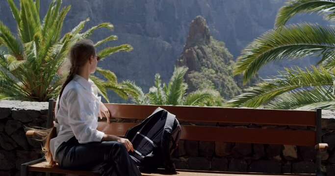 Masca Gorge and village, Tenerife. Beautiful young woman with backpack sits on a bench and enjoy the view. Slow motion
