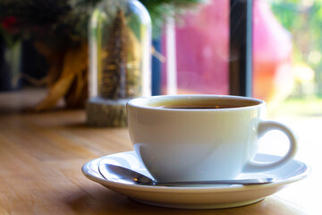 A white ceramic cup of hot black Americano coffee with light steam on top on a brown table in a coffee shop by the window. Drinks, relaxation, and holiday concept.