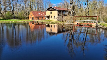 Obraz premium Water mill on the shore of lake and its reflection on the surface of the water in Latvian village of Garsenes in April 2022