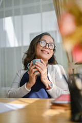 Obraz premium Pleased asian aged woman holding a mug of coffee, feeling fresh for start the day.