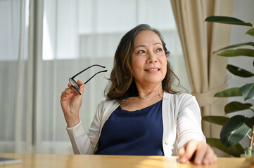 Successful asian aged woman holding her eyeglasses and looking through the window