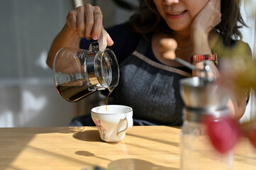 Cropped, aged woman poring hot coffee into a ceramic cup on the table.