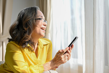 Cheerful asian aged woman holding, using smartphone and looking out the window