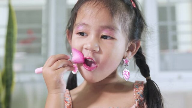 Asian adorable funny little girl making makeup her face she look in the mirror and cheek fluffy brush for powder, Learning activity to be woman, happy kid is beautiful make up with cosmetics toy