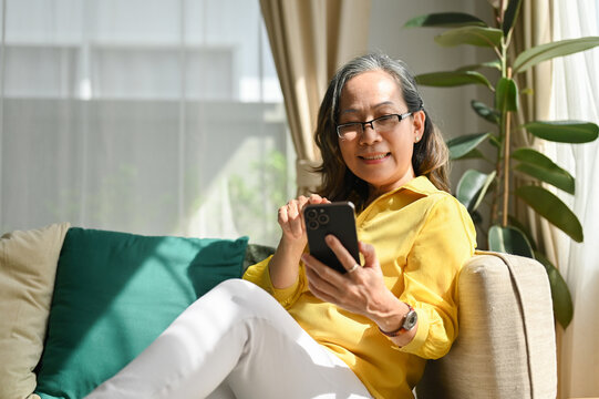 Relaxed asian retired aged woman sitting on comfortable couch and using cellphone