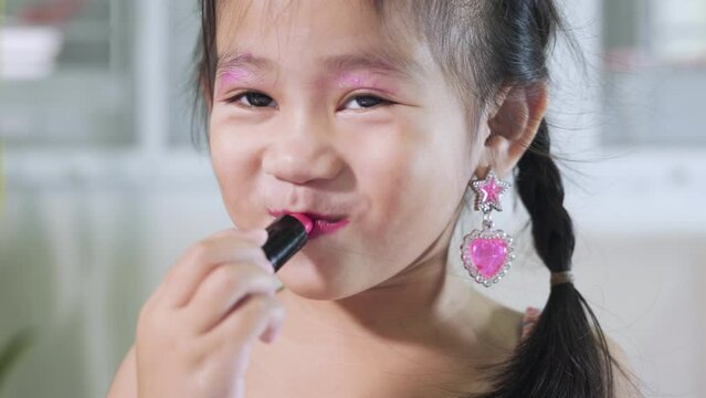 Asian adorable funny little girl making makeup her face she look in the mirror and applying red lipstick to mouth, Learning activity to be woman, happy kid is beautiful make up with cosmetics toy