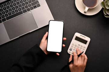 A female using smartphone and calculator in her modern black office desk. top view