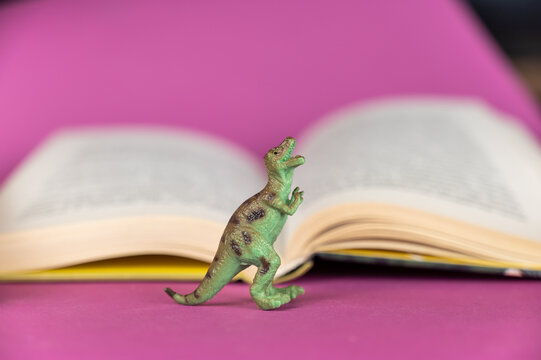 Dinosaur and open book against purple background. Green plastic miniature of an animal of prey. Hardcover book in the background. Selective focus.