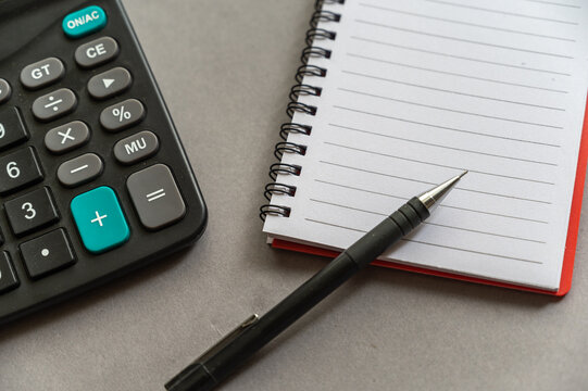 Calculator, pen, notepad lie on a gray surface. Open spring-loaded notebook with blank sheets. Close-up.