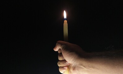 Hand tightly holding one lit burning white long wax candle light with fire flame dark black night...