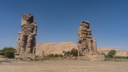 Giant sculptures of the colossi of Memnon against the blue sky and sand dunes. Huge statues of seated pharaohs are dilapidated. Egypt