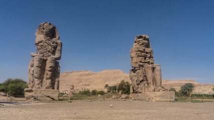 Giant sculptures of the colossi of Memnon against the blue sky and sand dunes. Huge statues of...