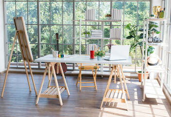 Wooden modern style working table with laptop notebook computer equipment with plant pot, colorful acrylic painting color, smartphone in glass windows workshop studio with canvas easel and bookshelf.