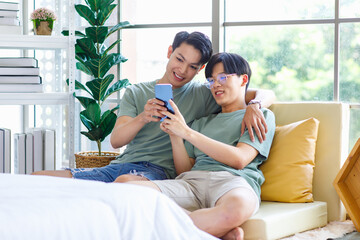 Two Asian young handsome pride male gay men lover couple partner sitting smiling together on cozy sofa in bedroom holding looking browsing surfing via smartphone cuddling hugging showing LGBTQ love
