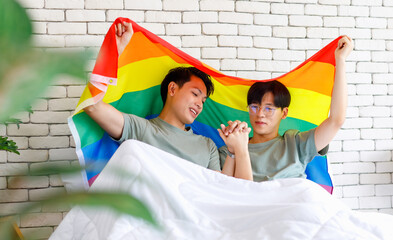 Two Asian happy cheerful romantic lovely teenager male gay men lover couple partner smiling lying down holding hands cuddling together on bed covering selves with colorful rainbow pride equality flag