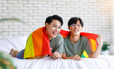 Portrait shot two Asian young lovely pride male gay men lover couple partner smiling laying down together in bed covered by rainbow sexual gender equality flag holding hands showing mini heart sign