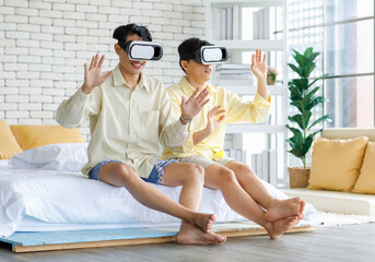 Two Asian young happy playful teenager male gay men lover couple partner sit on bed in modern bedroom wearing virtual reality VR goggles headset having fun playing online 3D simulator game together