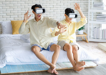 Two Asian young happy playful teenager male gay men lover couple partner sit on bed in modern bedroom wearing virtual reality VR goggles headset having fun playing online 3D simulator game together
