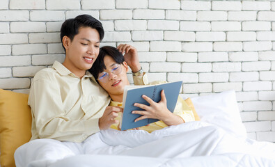 Two Asian young happy romantic teenager male gay men lover couple partner laying down under blanket on bed in bedroom leaning at brick wall background cuddling hugging together while reading novel