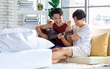Two Asian young happy romantic lovely teenager male gay men lover couple partner sitting smiling on cozy sofa in bedroom cuddling hugging together while husband playing acoustic guitar and sing song