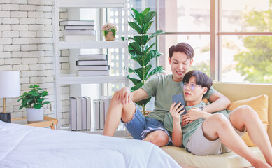 Fototapeta premium Two Asian young handsome pride male gay men lover couple partner sitting smiling laying down together on cozy sofa in bedroom cuddling hugging watching entertainment media streaming on smartphone