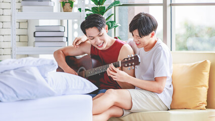 Fototapeta premium Two Asian young happy romantic lovely teenager male gay men lover couple partner sitting smiling on cozy sofa in bedroom cuddling hugging together while husband playing acoustic guitar and sing song
