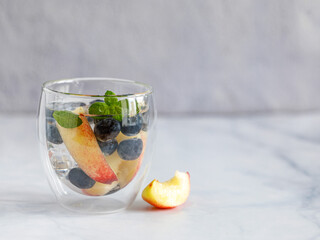 Blueberry and peach infused water, cocktail, lemonade or tea in double glass. Summer iced cold drink with blueberry, ice, peach and mint on grey marble background, copy space, side view
