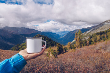 Hand holding enamel white mug mockup with forest and mountains valley background. Trekking merchandise and camping gear. Stock wildwood photo with white metal cup. Rustic product template.