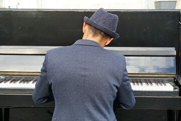 Rücken von jungem Pianist vor schwarzem Klavier  gekleidet mit blau-grauem Blazer und Hut bei der...