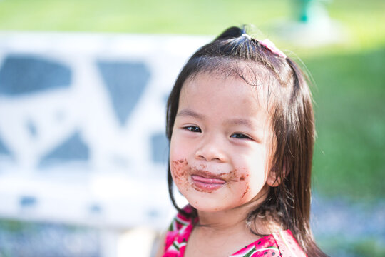 Candid image baby 3-4 years old. Cute little Asian girl is licking chocolate smeared all over her lips and cheeks. Face children enjoy dessert in afternoon. Summer or spring season. Child sweet smile.