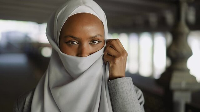 Cheerful African-American woman covers face with hijab on waterfront closeup slow motion. Advanced Muslimah shows traditional accessory in city